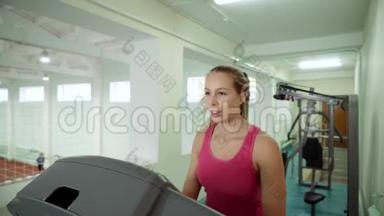 漂亮的金发女郎在健身房在跑步机上做运动。 那个女孩沿着跑步机跑步。